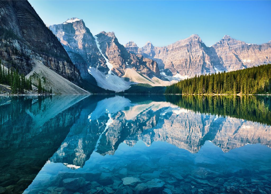 Clear turquoise lake reflecting jagged, snow-capped mountains and pine forests under a bright blue sky, with rocks visible beneath the water’s surface in the foreground—a breathtaking landscape that captures the natural beauty awaiting those who pursue Cursos profesionales Canadá. Live and Work In Australia